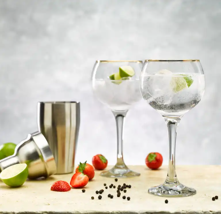Two gin and tonics in goblets, a cocktail shaker with limes, strawberries and pepp ercornson the counter 